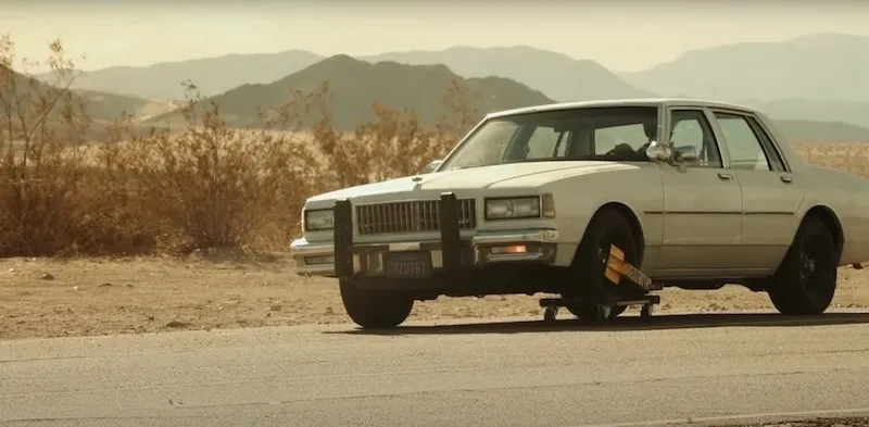 A screen capture from paradox bullets by Tom Sachs. It shows a white caprice with a wheel lock on the front left tyre. A movers dolly has been used to circumvent the lock.